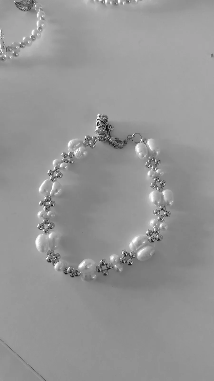 A black-and-white photo shows a handmade pearl and silver bead bracelet with a decorative clasp, resting on a plain surface…