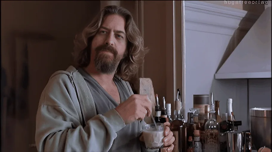 The image shows a man with long hair and a beard, wearing a light-colored robe, standing in a kitchen holding a glass and a…