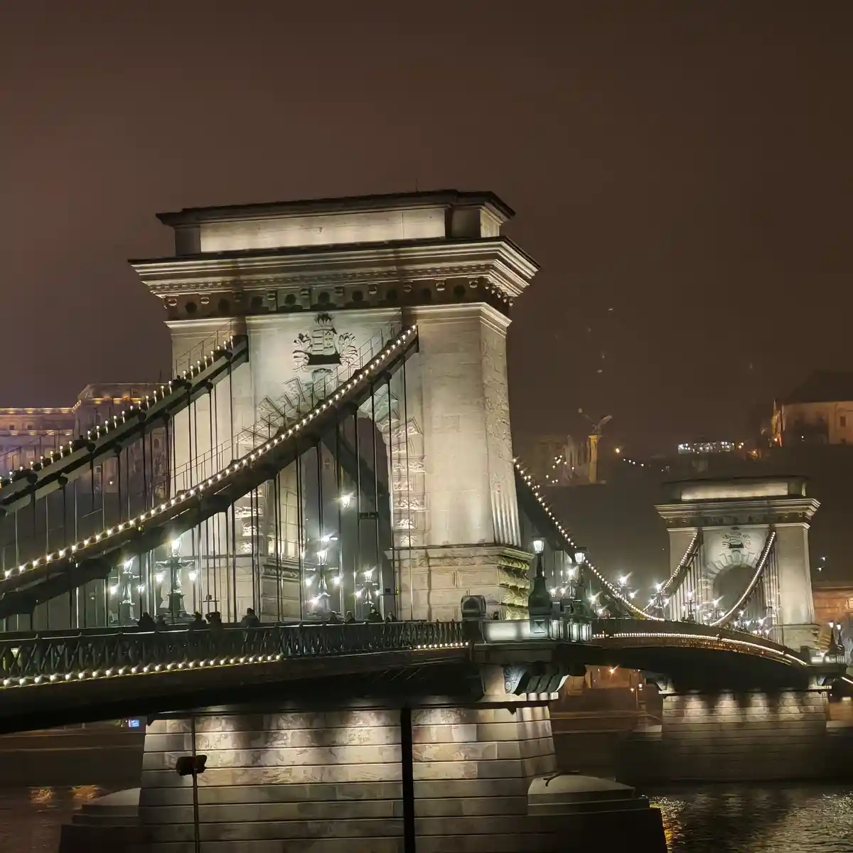 A nighttime photo of the illuminated Chain Bridge in Budapest, showing its stone towers, suspension cables, and ornate…