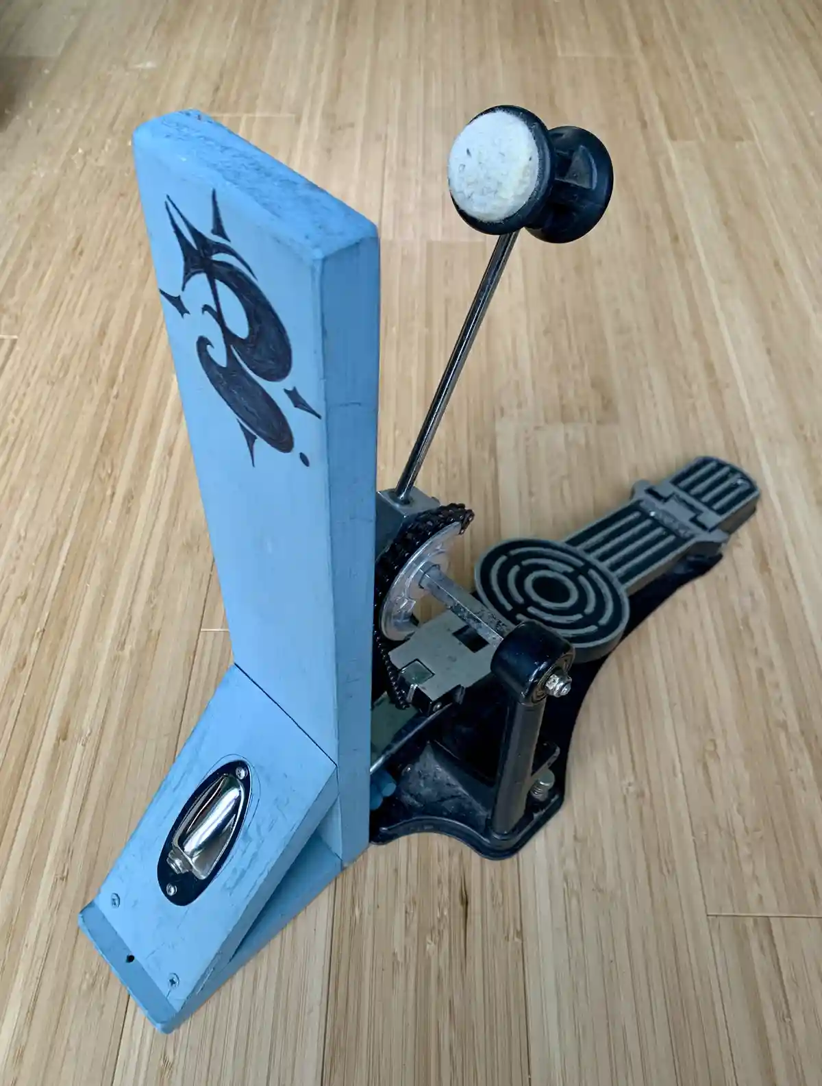 A close-up photo of a blue-painted wooden object with a black tribal-style design, attached to a black metal pedal mechanism…