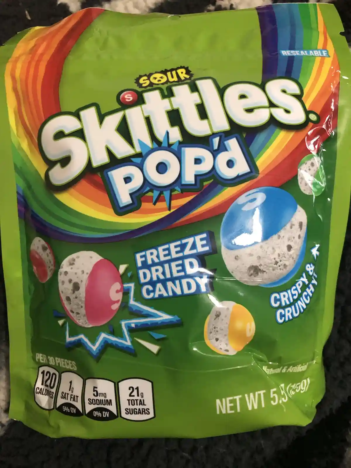 A close-up of a green bag of Sour Skittles Pop'd candy, featuring images of freeze-dried, half-colored, half-white spherical…