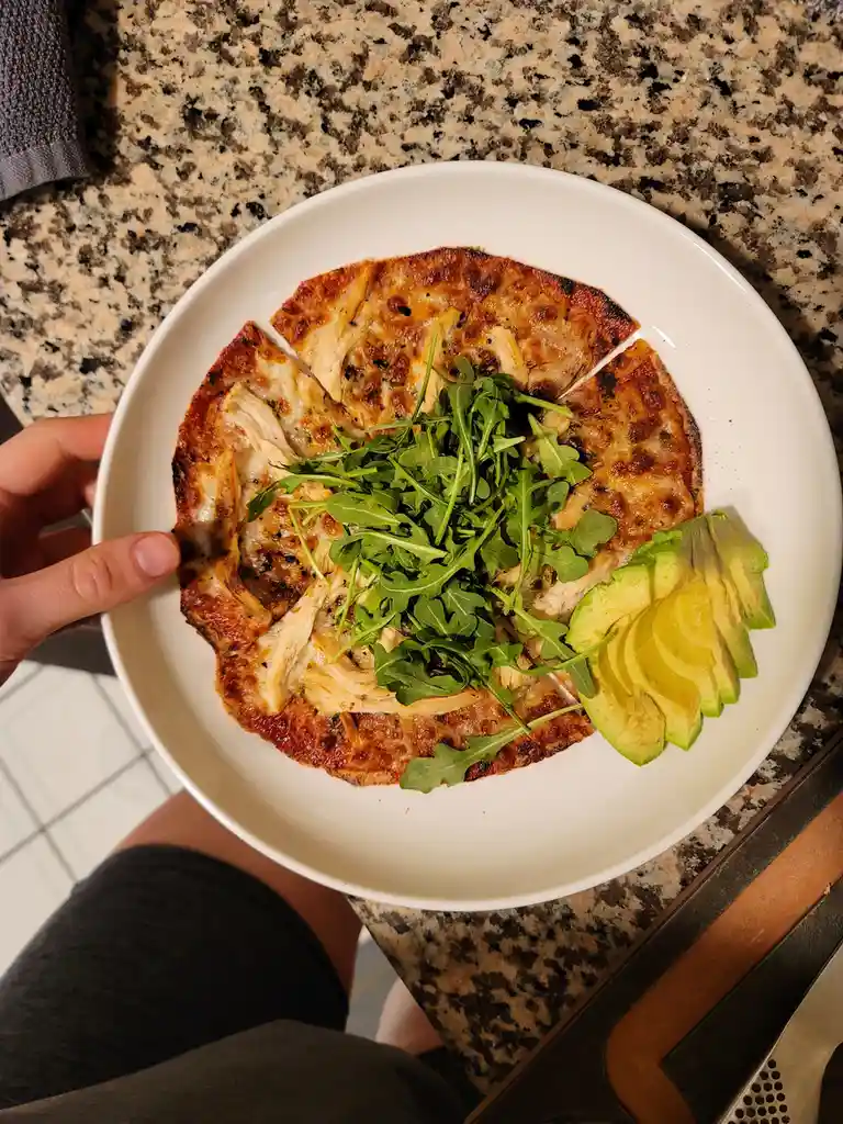 A person holds a white plate with a thin, sliced flatbread topped with shredded chicken, melted cheese, and a pile of fresh…