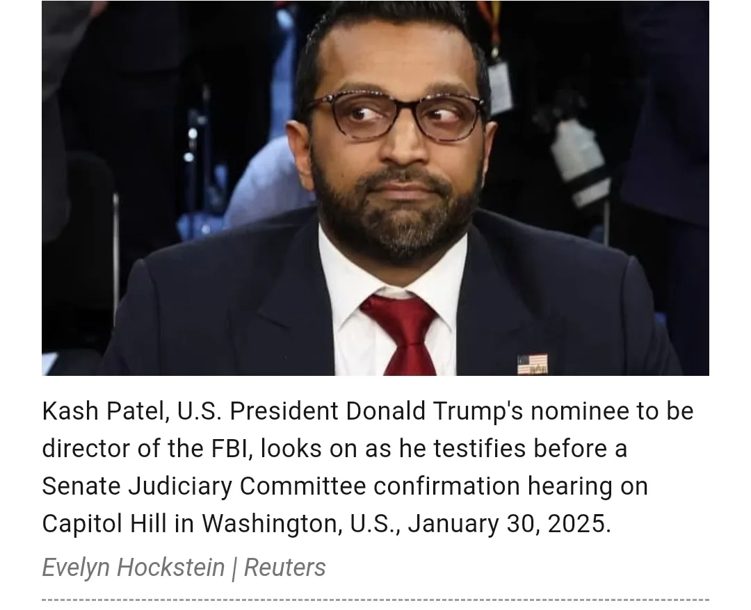 A man with glasses and a beard, wearing a dark suit, white shirt, and red tie, looks sideways with a skeptical expression…