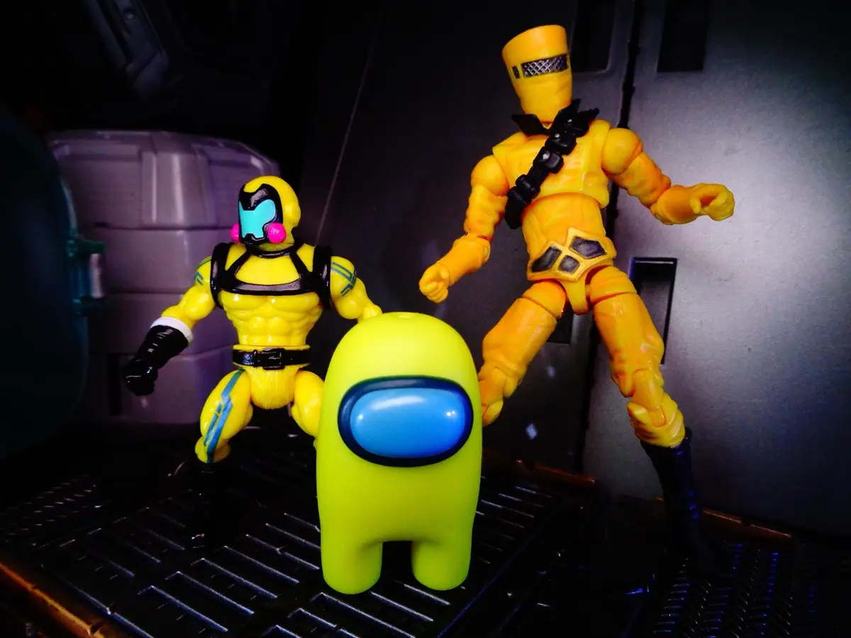 Two yellow action figures, resembling characters from the game 'Among Us', stand on either side of a lime-green 'Among Us'…