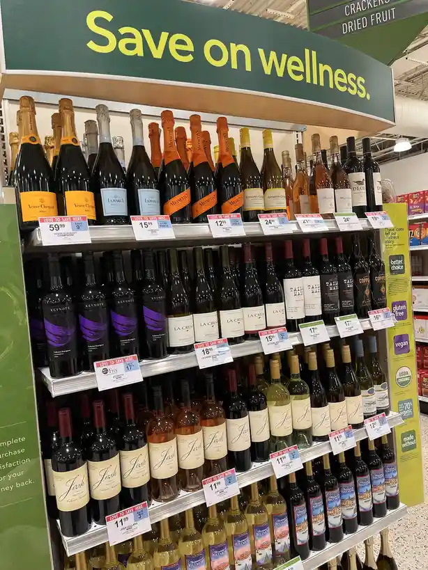 A supermarket aisle displays shelves of wine bottles under a green sign that reads 'Save on wellness.' The products include…