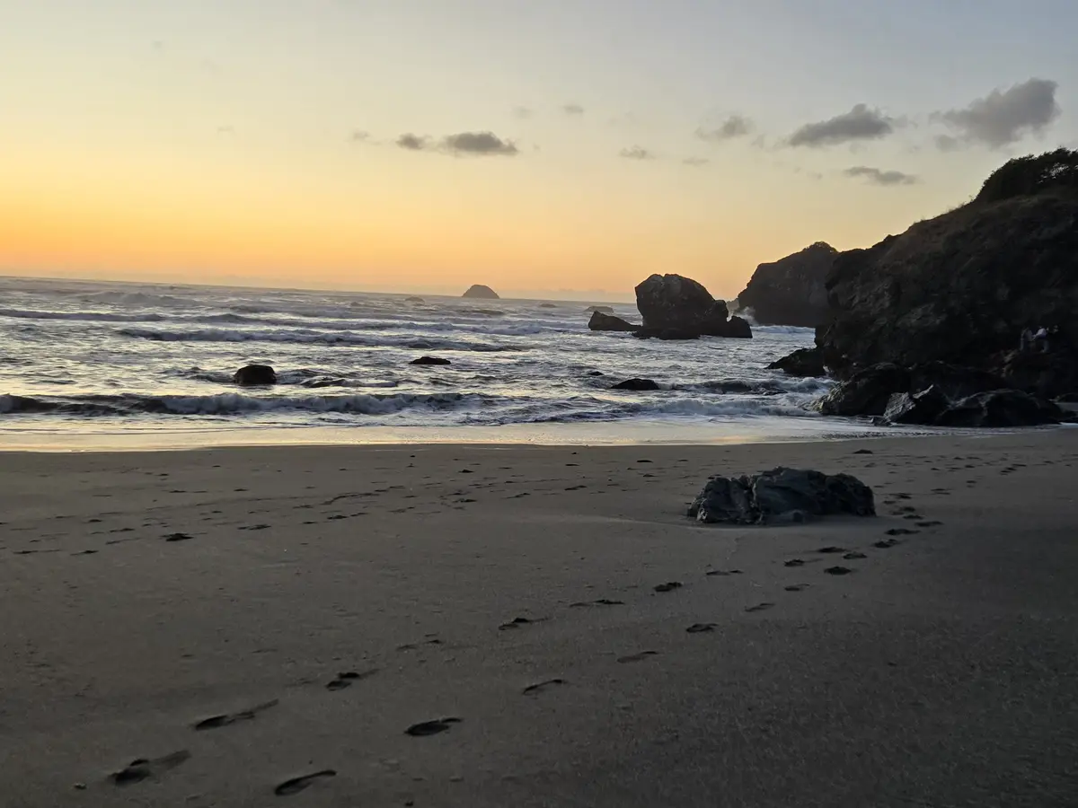 A serene beach sunset photo shows footprints leading toward the ocean, with waves crashing against rocks and a cliffside in…