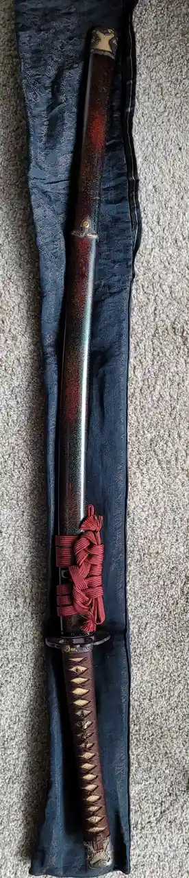 A traditional Japanese katana sword with a dark, textured blade and red braided tsuka (handle) is displayed vertically…