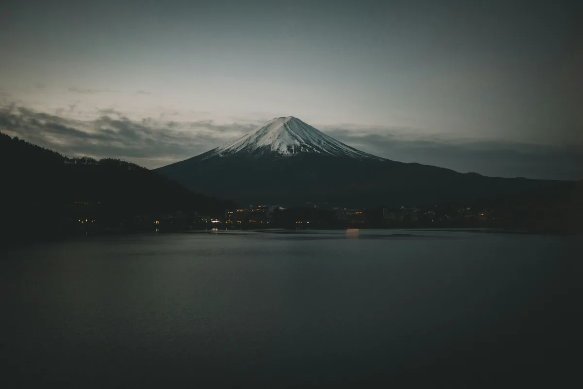 A serene, dimly lit photograph of Mount Fuji with snow-capped peaks, viewed across a calm lake at dusk or dawn. Silhouetted…