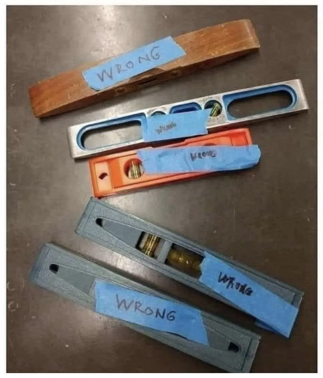 Five spirit levels of varying sizes and colors lie on a metal surface, each with a blue tape label handwritten with the word…