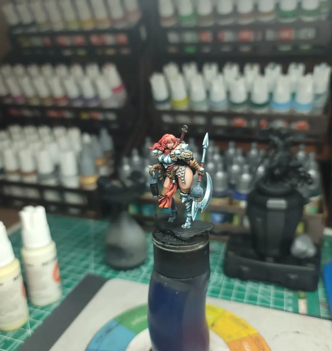 A painted fantasy miniature warrior figure with red hair, armor, and weapons stands on a black base atop a blue water…