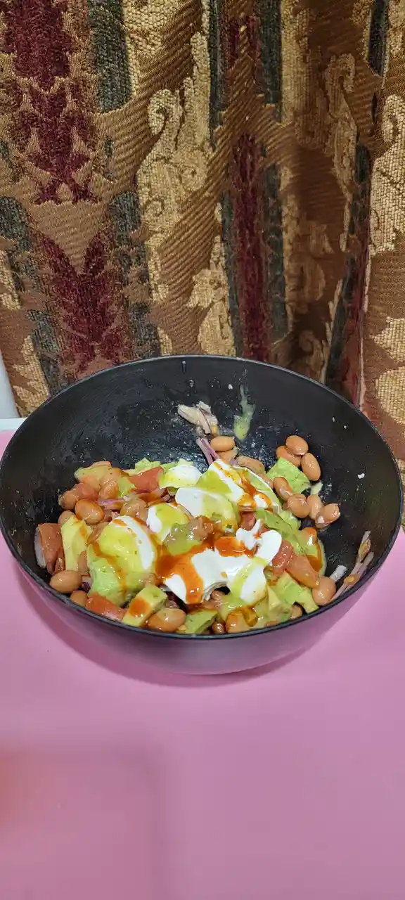 A black bowl filled with a colorful salad of beans, avocado, tomato, red onion, and white sauce drizzled with orange hot…
