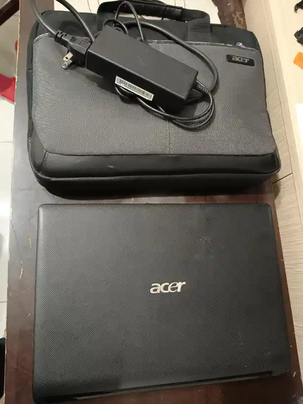 A black Acer laptop sits closed on a wooden surface, with its matching black textured laptop bag placed above it. A black AC…