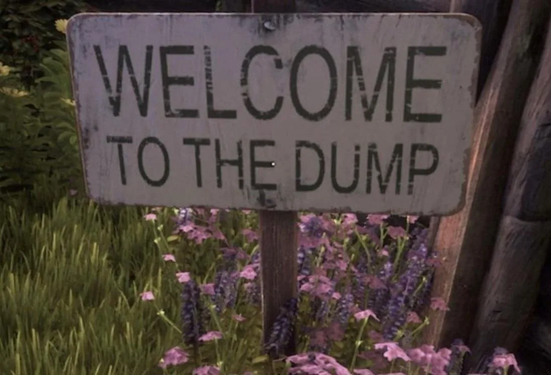 A weathered, white rectangular sign with black stenciled text reading 'WELCOME TO THE DUMP' is mounted on a wooden post. The…