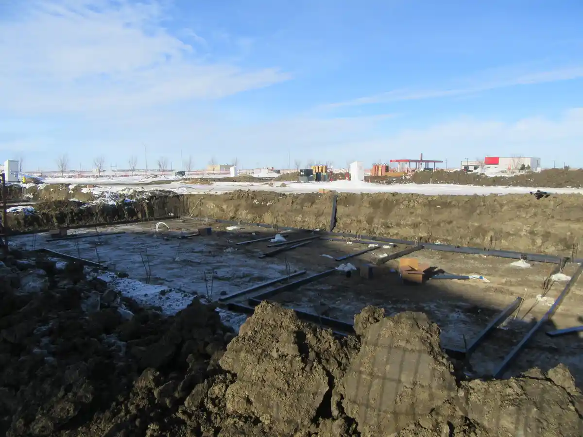 A photo of a construction site with exposed steel beams and dirt, partially covered in snow under a blue sky, overlaid with…