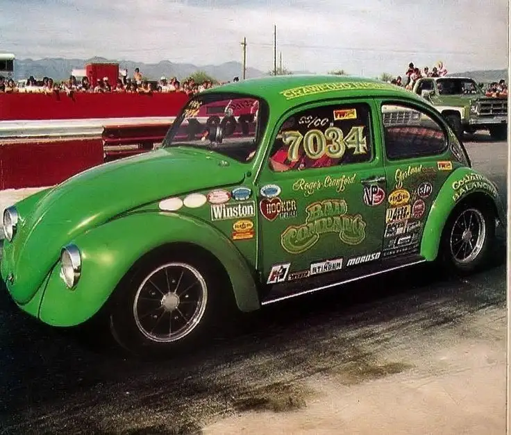 A bright green, heavily modified Volkswagen Beetle race car, number 7034, is shown on a drag strip with spectators in the…