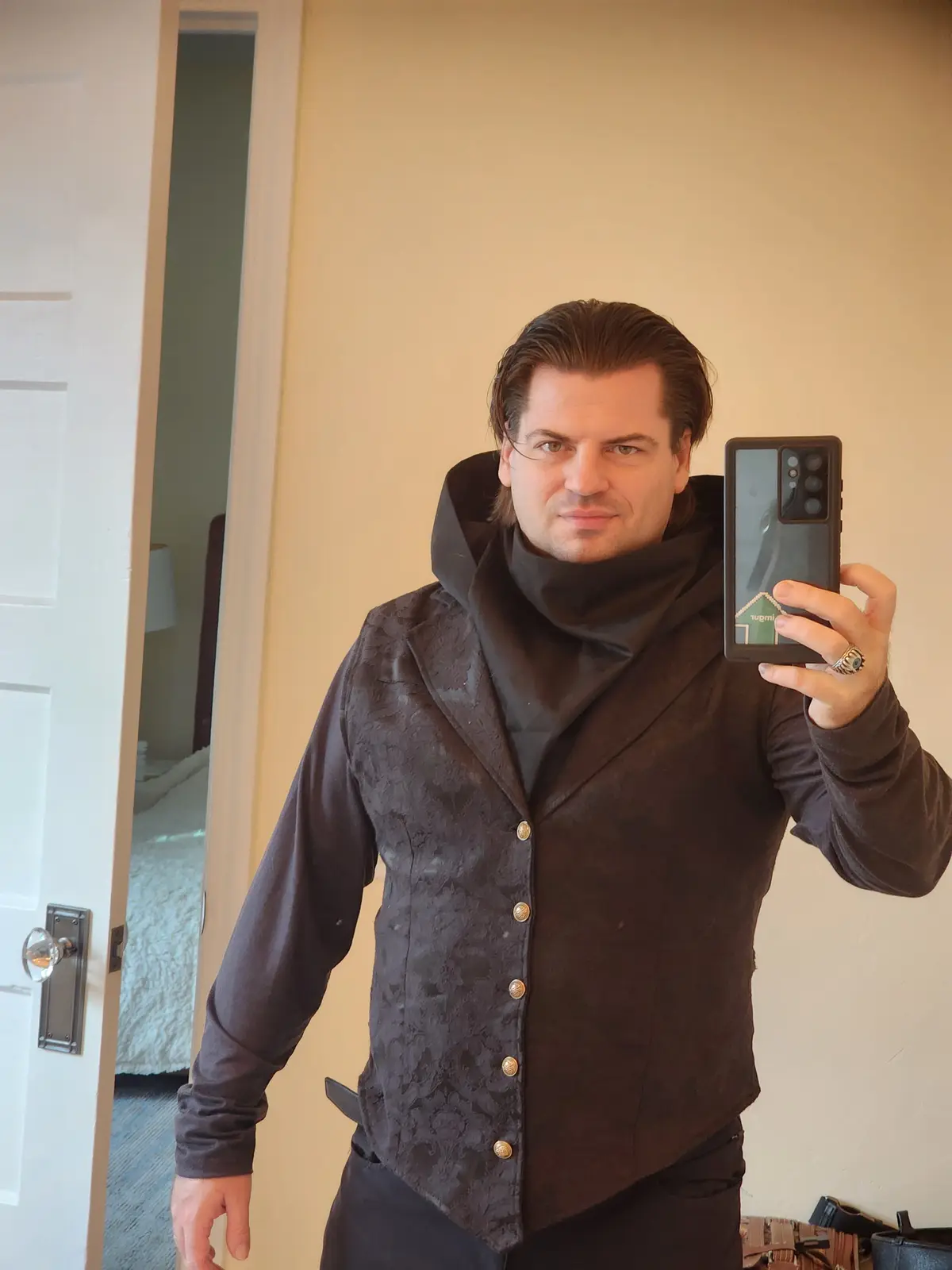 A man with slicked-back hair takes a mirror selfie in a dark, textured, button-up jacket with a high collar, holding a…