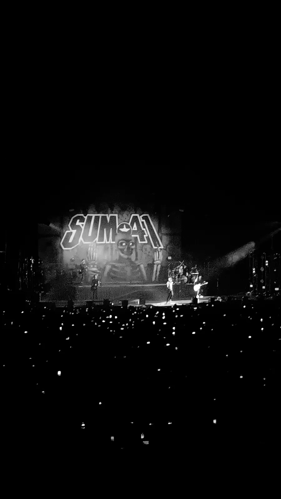 A black and white photo of a live concert with the band Sum 41 performing on stage, their logo prominently displayed behind…
