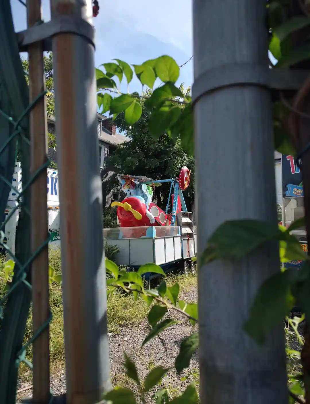 A weathered, colorful toy airplane ride sits abandoned on a trailer behind a chain-link fence, partially obscured by green…