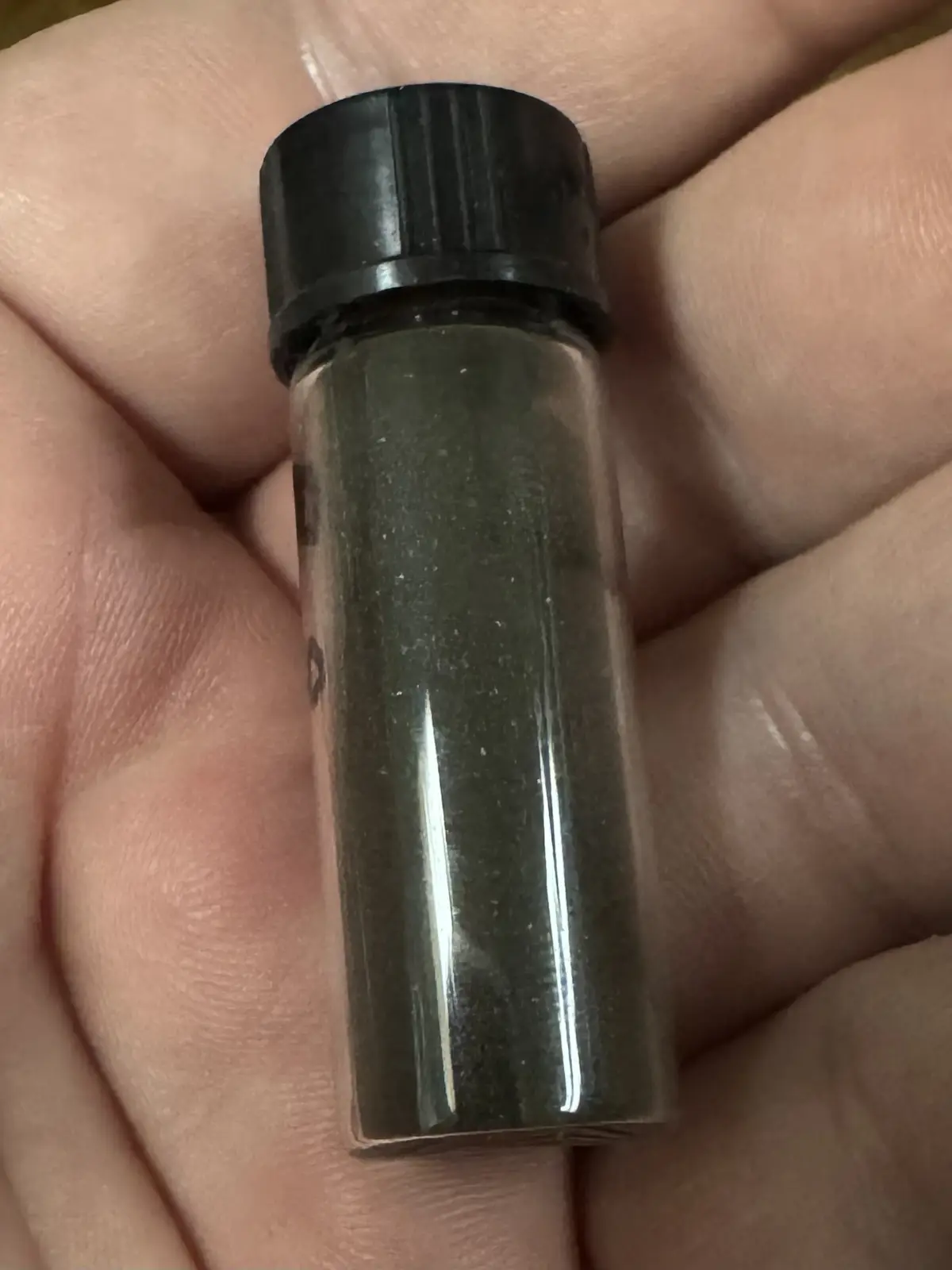 A hand holds a small, clear plastic vial with a black cap, filled with a dark green granular substance. The overlaid text…