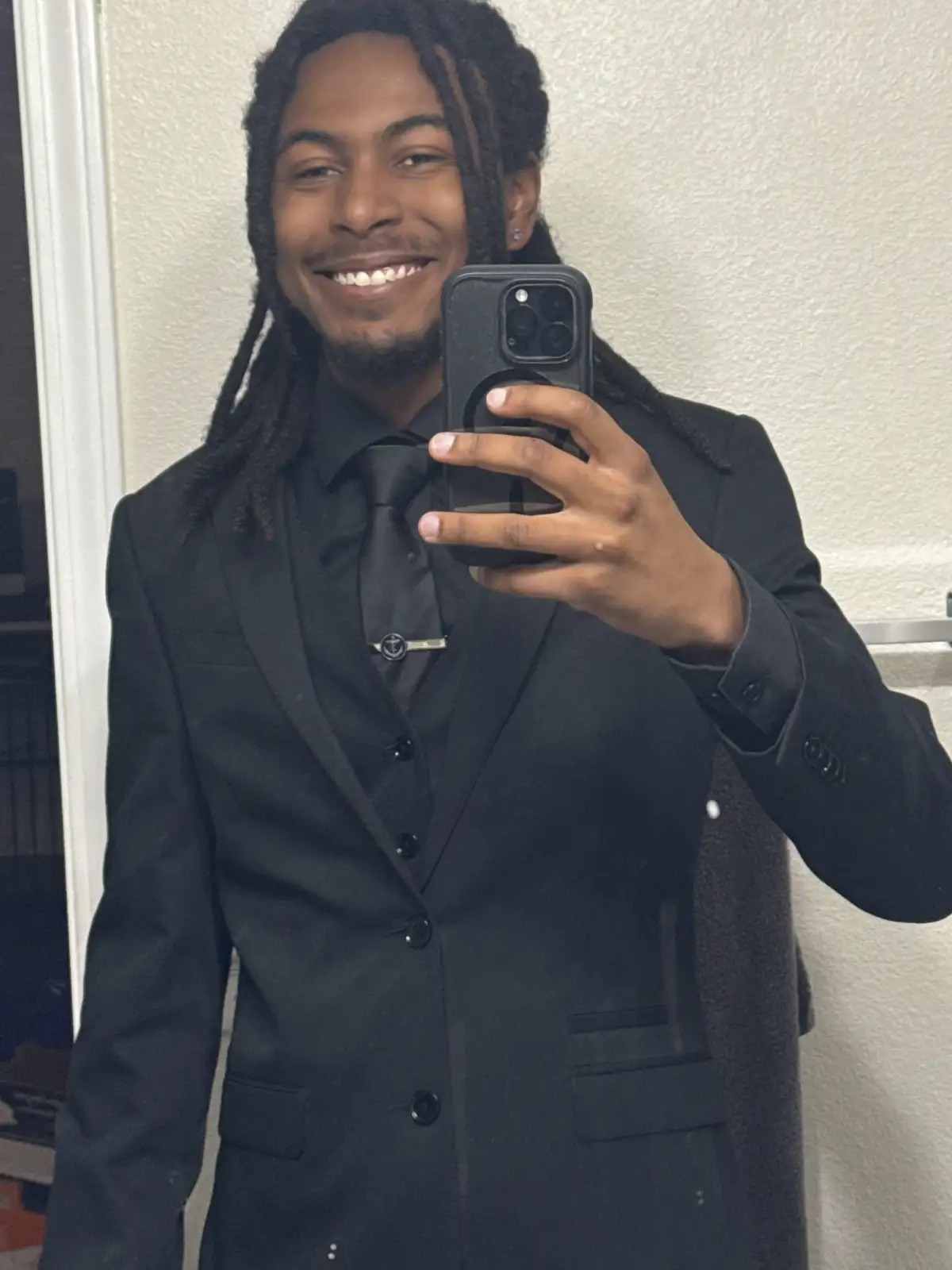 A smiling young man with dreadlocks takes a mirror selfie while wearing a sharp black suit, shirt, and tie. He holds a black…