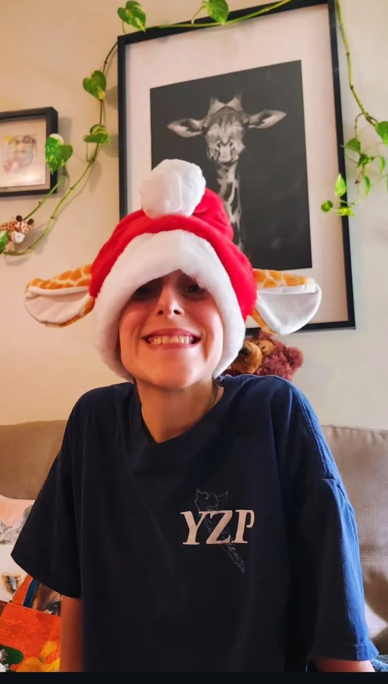 A person wearing a red and white Santa hat with giraffe ears and a pom-pom, smiling while seated on a couch. Behind them is…