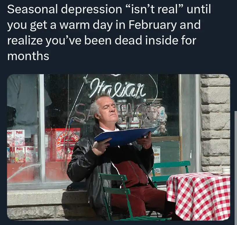A man in a black leather jacket sits outside an Italian sausage shop, looking upward with a weary expression while holding a…