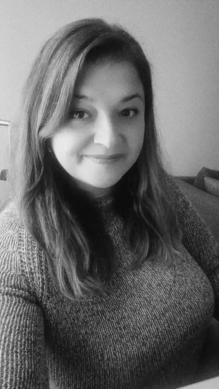 A black-and-white photo of a woman with long hair wearing a textured sweater, smiling slightly while looking at the camera…
