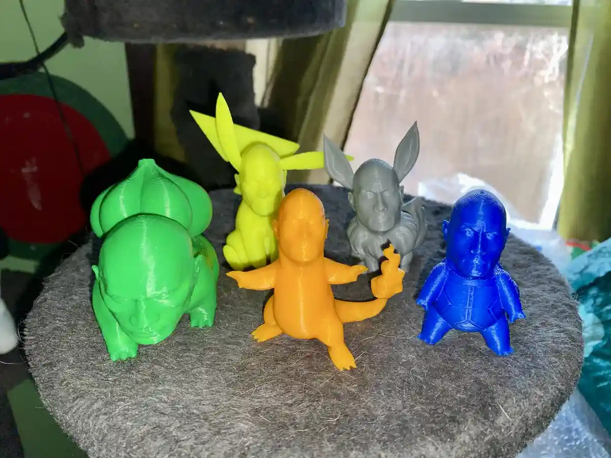 Four 3D-printed Pokémon figures—Bulbasaur, Pikachu, Eevee, and Squirtle—are arranged on a gray, fuzzy surface, each with a…