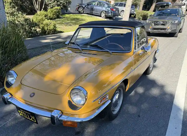 A gold Fiat 124 Spider convertible is parked on a residential street, its California license plate reading 'STILL25'. Other…
