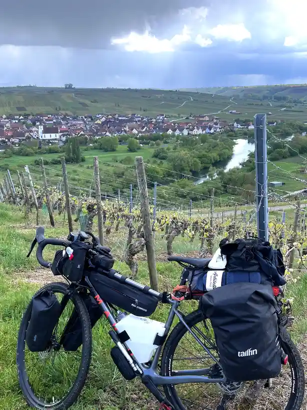 A heavily loaded touring bicycle with panniers and gear is parked in a vineyard overlooking a scenic valley town and river…
