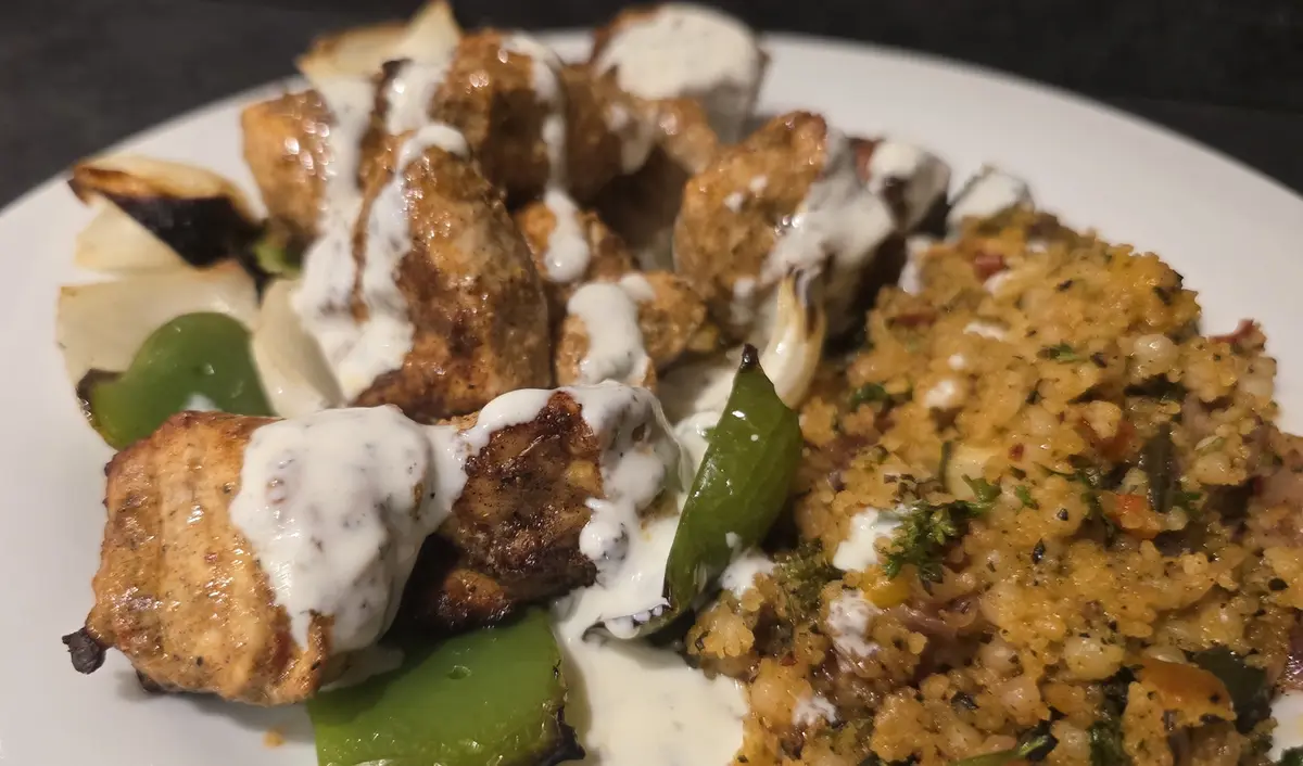 A close-up photo of a plated meal featuring grilled chicken pieces generously drizzled with a white sauce, accompanied by…
