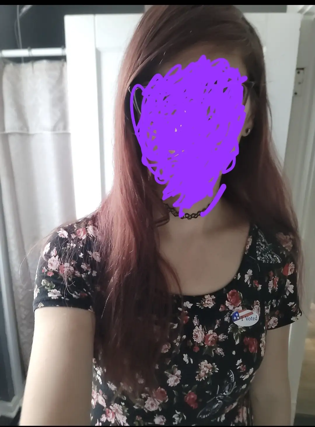 A person with long reddish-brown hair wears a black floral-patterned shirt and a choker necklace. Their face is obscured by…