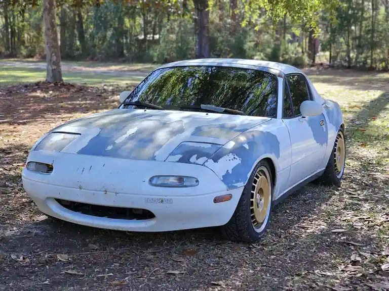 A white Mazda Miata sports car with peeling paint and dirt is parked in a wooded area with fallen leaves on the ground. The…