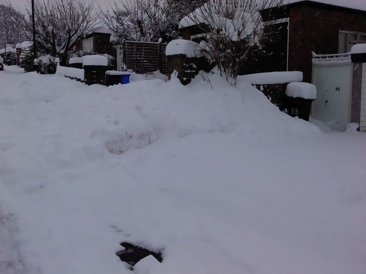 A snowy residential street with houses, fences, and bushes completely buried under deep snow. A single dark footprint is…