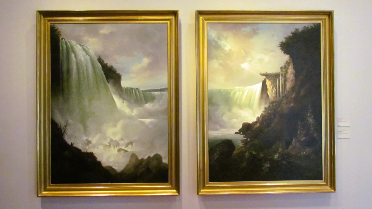 Two identical gold-framed paintings of Niagara Falls hang side-by-side on a wall, each depicting the same waterfall scene…