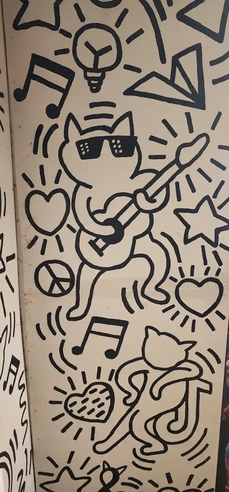 A black-and-white mural features a stylized cat wearing sunglasses and playing a guitar, surrounded by symbols like hearts…