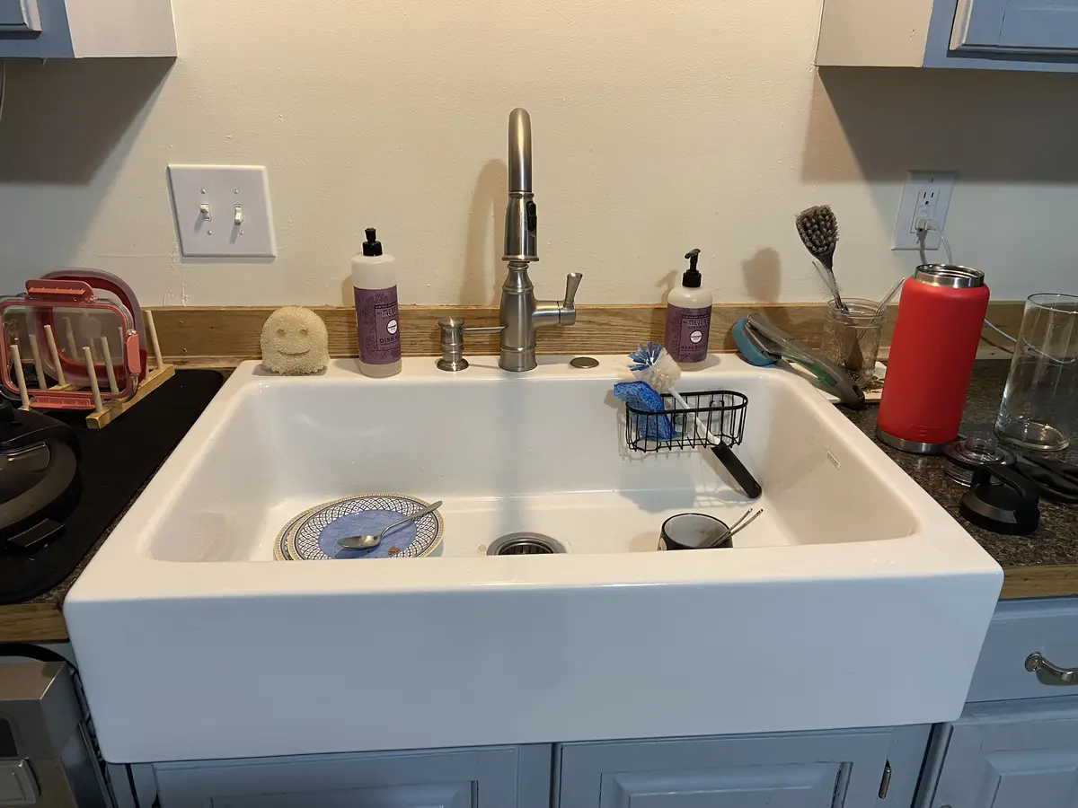A white farmhouse-style kitchen sink with a single plate and spoon inside, surrounded by dish soap, sponges, and cleaning…