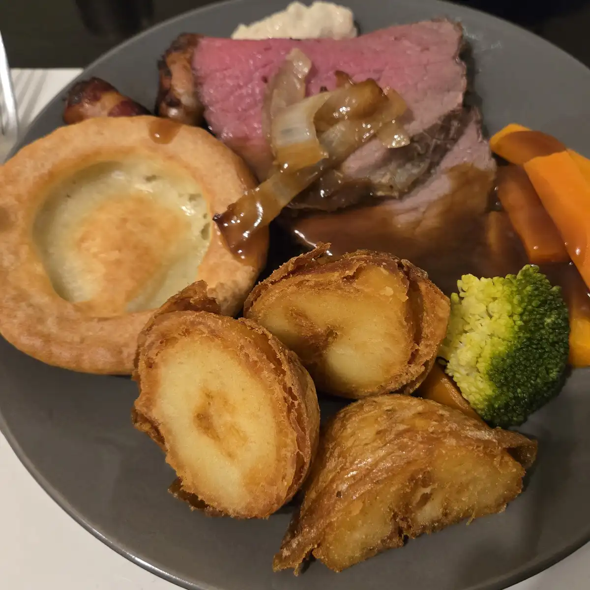 A close-up photo of a traditional roast dinner on a grey plate, featuring a slice of medium-rare beef with gravy and…