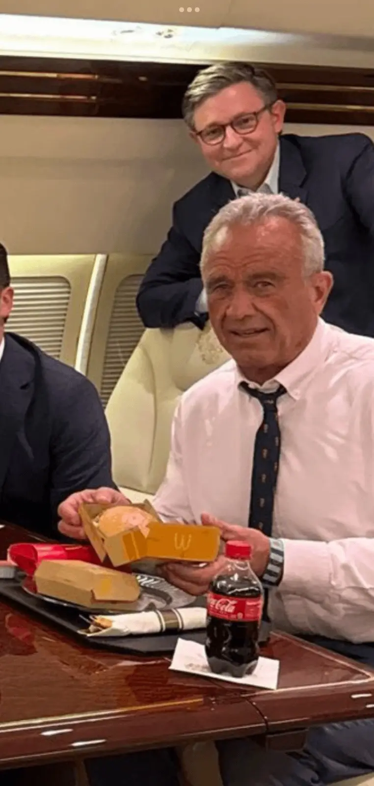 An older man in a white shirt and tie sits at a polished wooden table on a private jet, holding open a McDonald's burger box…