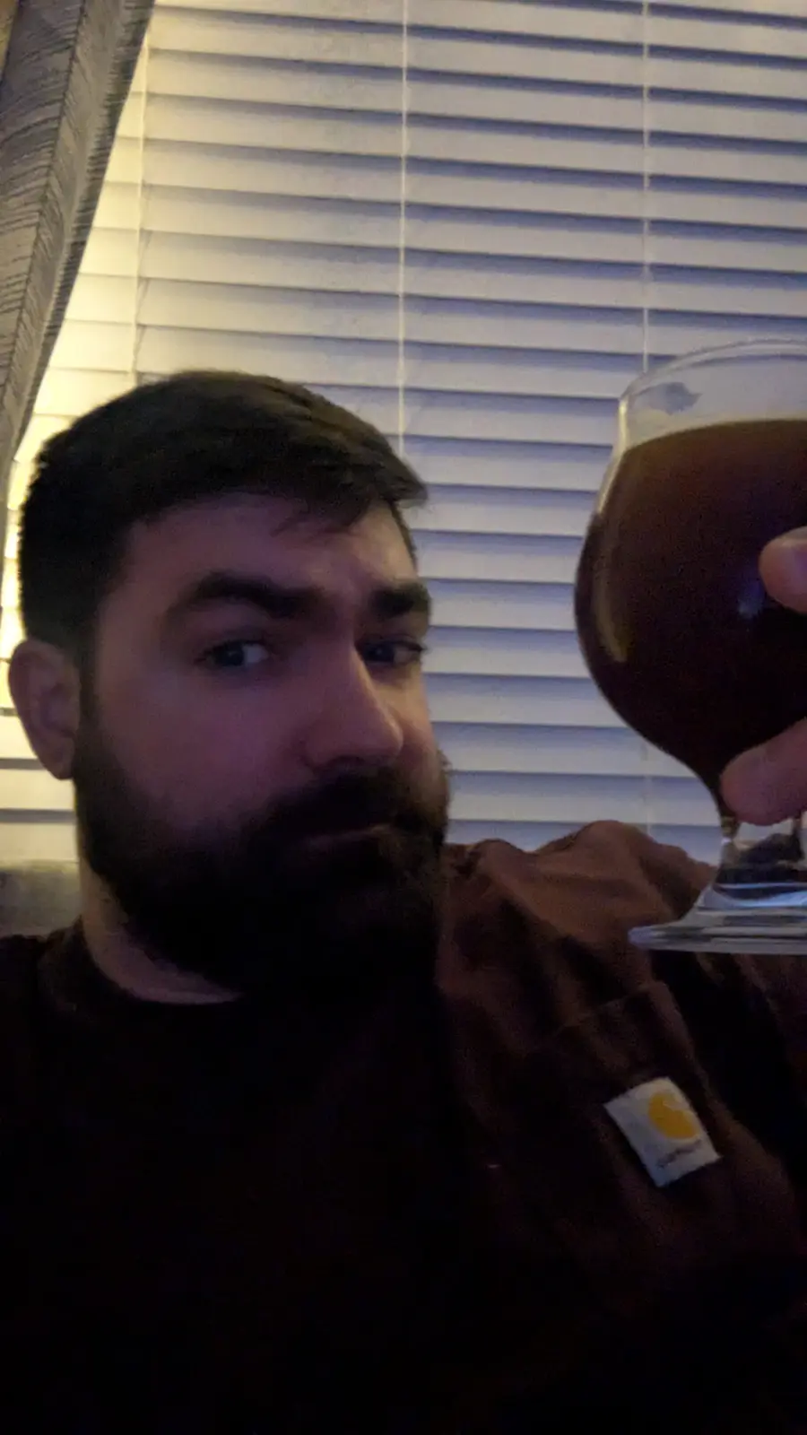 A man with a beard and dark hair, wearing a brown shirt with a Carhartt logo, holds up a glass of dark beer while looking…