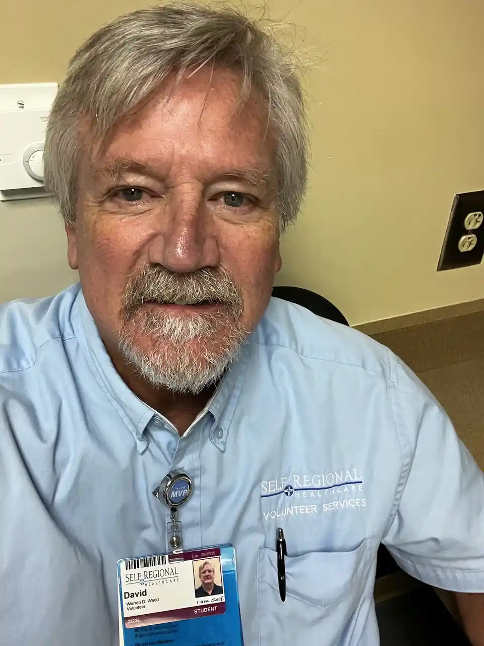 A man with gray hair and a mustache wears a light blue 'Self Regional Healthcare Volunteer Services' shirt and a name badge…