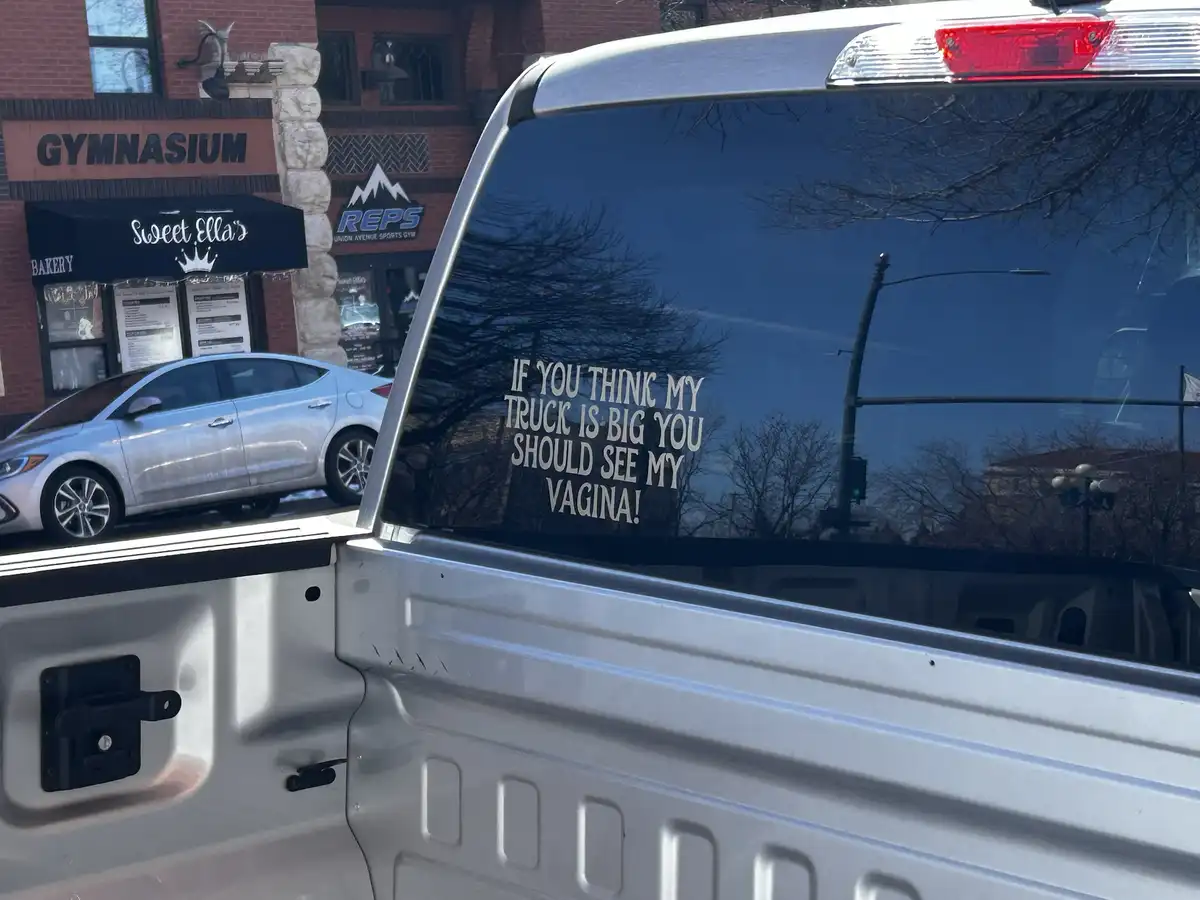 A silver pickup truck's rear window displays a white decal with the text 'IF YOU THINK MY TRUCK IS BIG YOU SHOULD SEE MY…