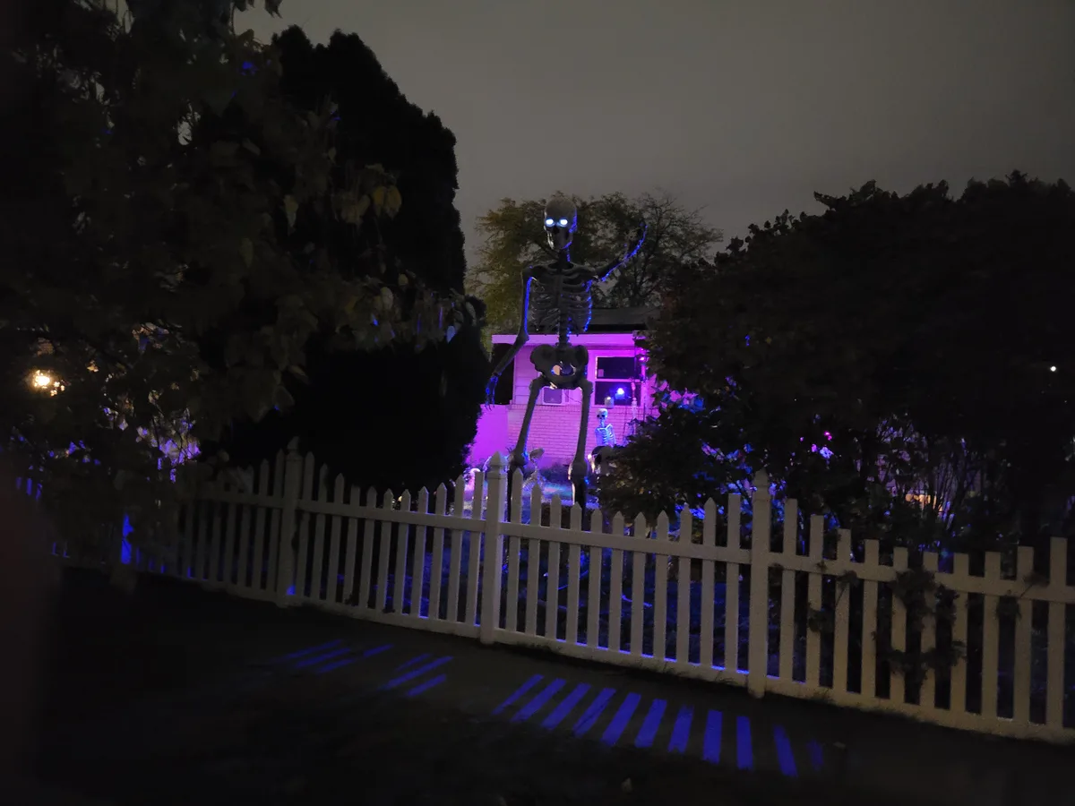A large, glowing skeleton decoration stands behind a white picket fence at night, illuminated by purple and blue lights. The…