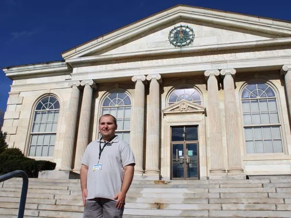 A man in a grey polo shirt and lanyard stands smiling on the steps of a large, classical-style stone building with columns…