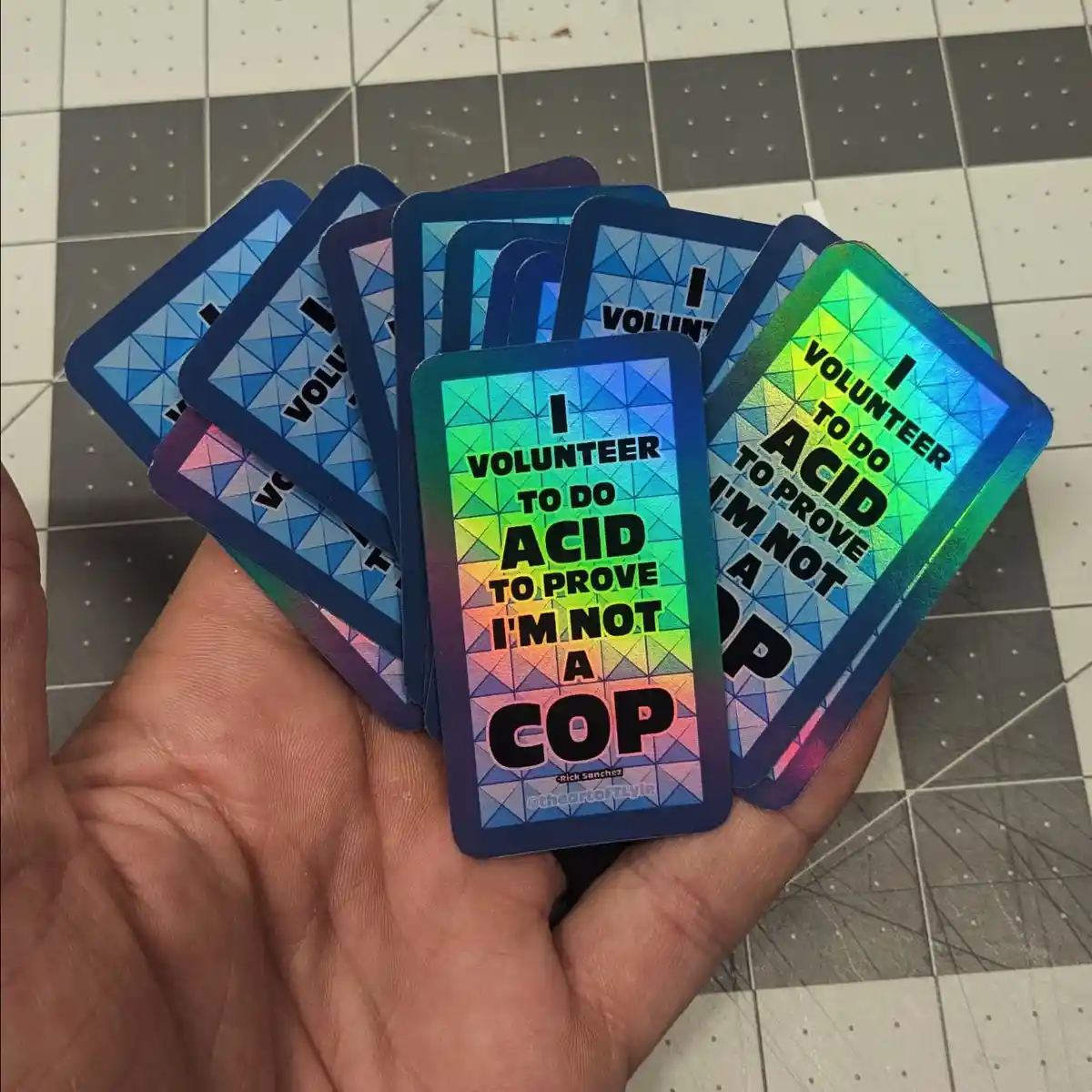 A hand holds a fanned-out stack of holographic cards, each featuring the text 'I VOLUNTEER TO DO ACID TO PROVE I'M NOT A…