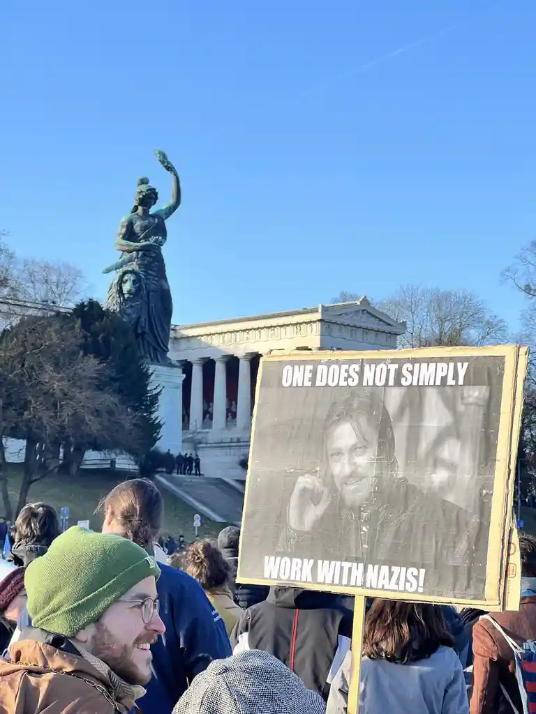 A protest scene with a crowd gathered outdoors under a clear blue sky, featuring a large bronze statue of a woman holding a…