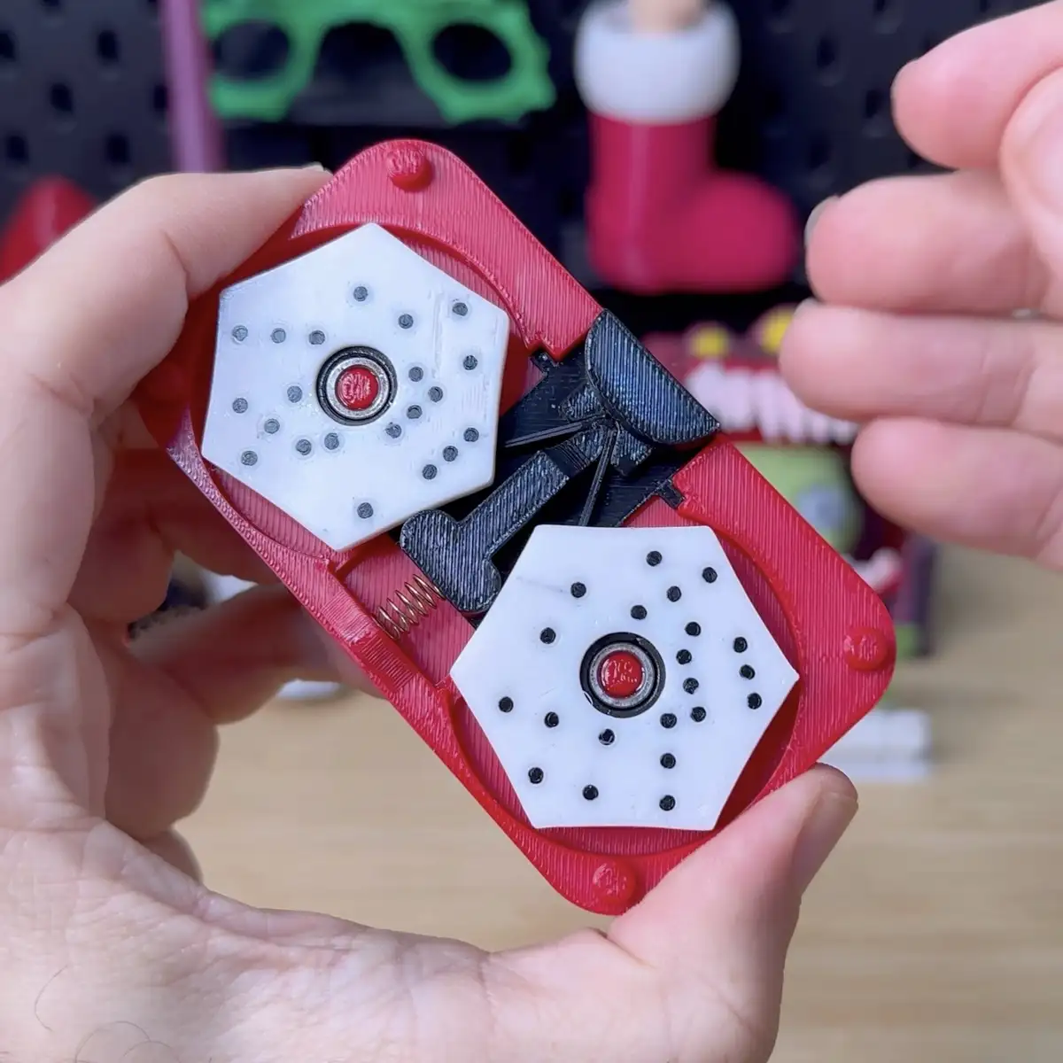 A person holds a red 3D-printed fidget spinner with two white hexagonal weighted ends and black plastic arms, revealing its…