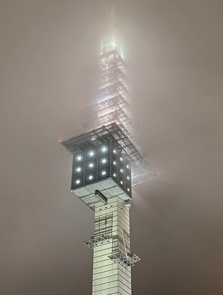 A low-angle nighttime photo shows a tall, illuminated tower partially obscured by thick fog, with lights visible on its…
