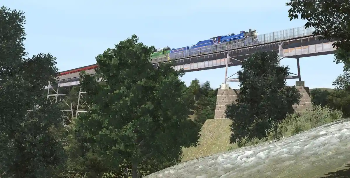 A blue steam locomotive pulls a train across a metal bridge over a river, with green trees and a grassy embankment below…