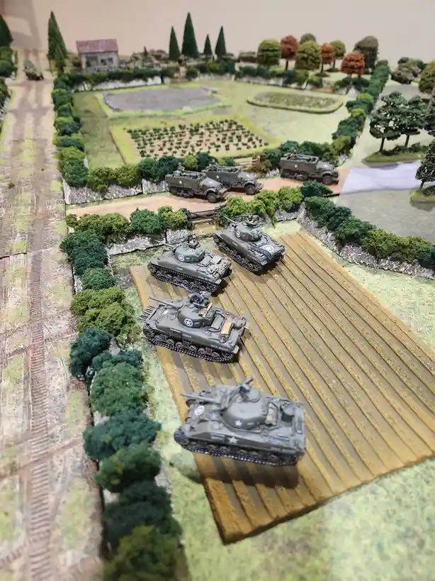 A detailed miniature diorama depicts a World War II battlefield with several olive-drab American military tanks and armored…